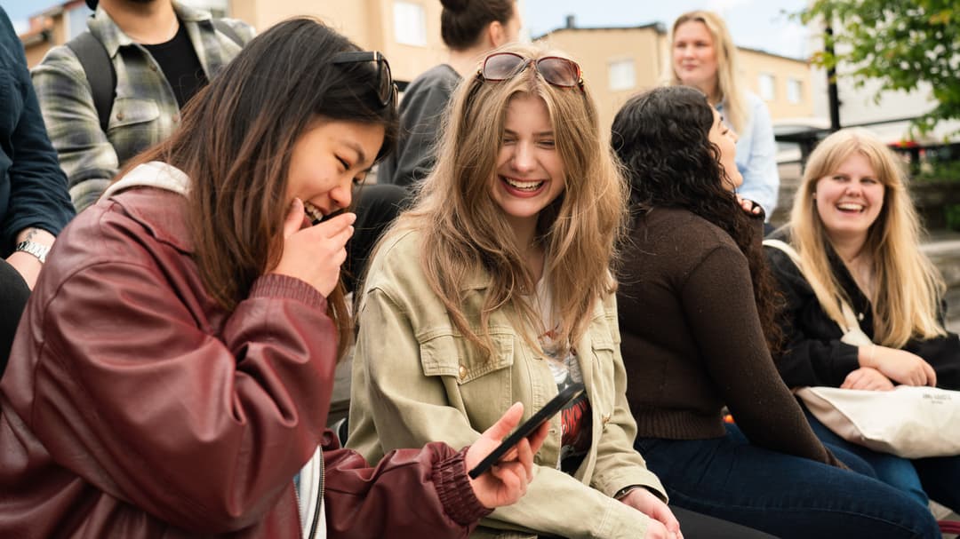 Två kvinnliga studenter tittar i en mobiltelefon. De sitter på en trappa på campus tillsammans med fler studenter.