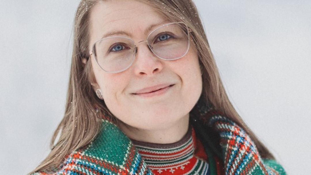 Woman with glasses and dark blonde hair with a chequered shawl over her shoulders looks at the camera.