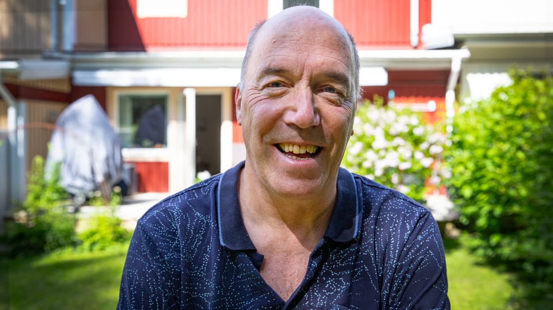 En man står med ryggen mot ett hus med lummig trädgård.
