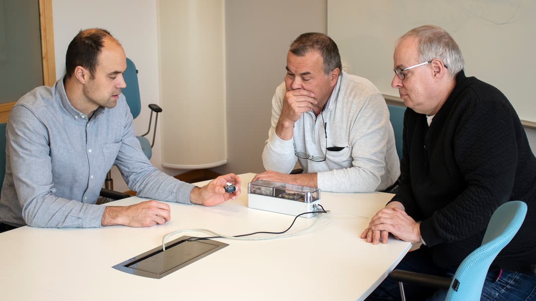 Tre män sitter vid ett bord och diskuterar en prototyp som ligger på bordet.