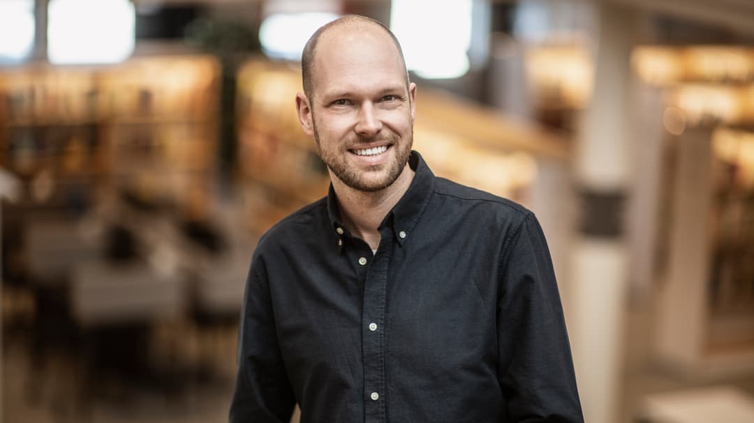 En man i svart skjorta med vita knappar ler mot kameran