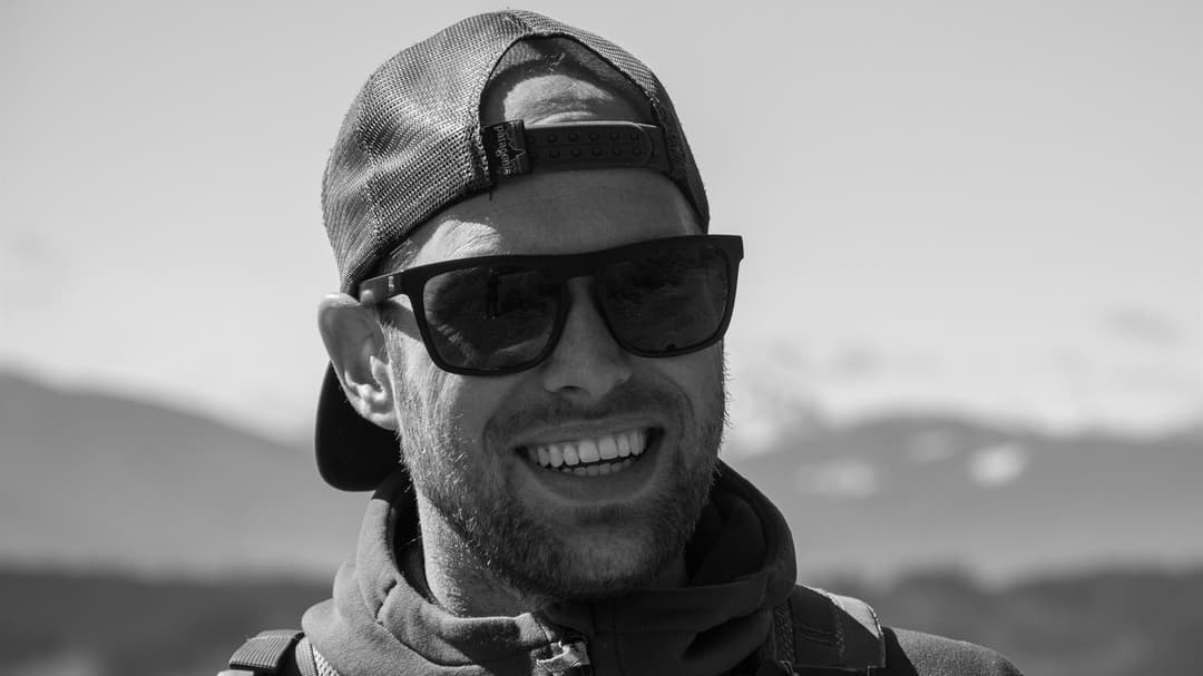 A black and white photo of young man standing in natural landscape with mountains in the background. He wears sunglasses and a cap back and front.