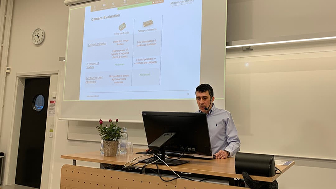 Man standing in front of computer presenting his thesis that can be seen on a screen in the background.