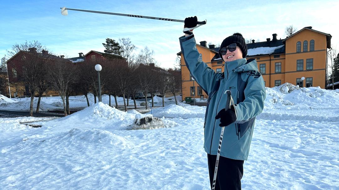 Ung man med stavar i händerna står på en snötäckt plan och sträcker upp ena handen i luften och ler