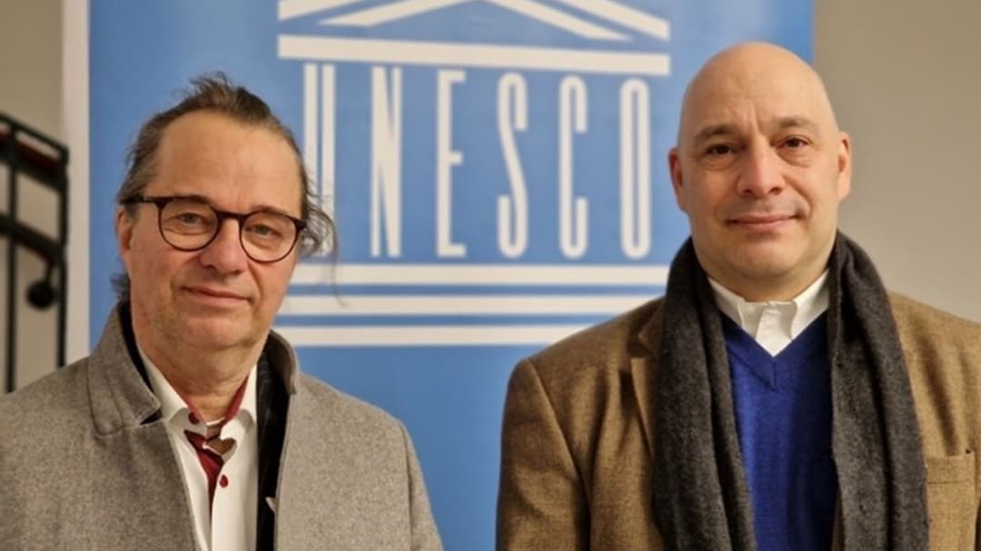 Två män klädda i rockar står framför en blå skylt med UNESCO-logotyp 