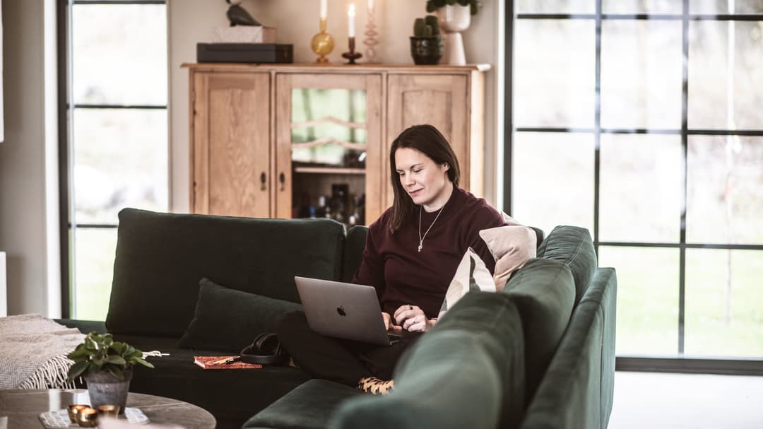 En kvinna sitter i soffan och jobbar med sin dator i knät. 