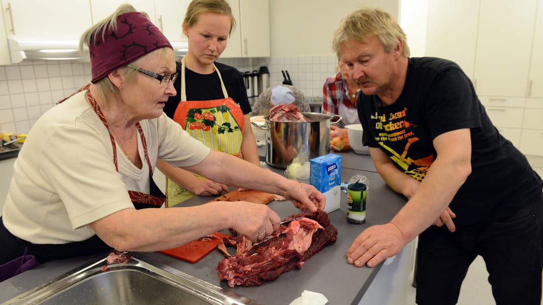 En äldre kvinna visar en ung kvinna och en man hur man tillagar renkött.