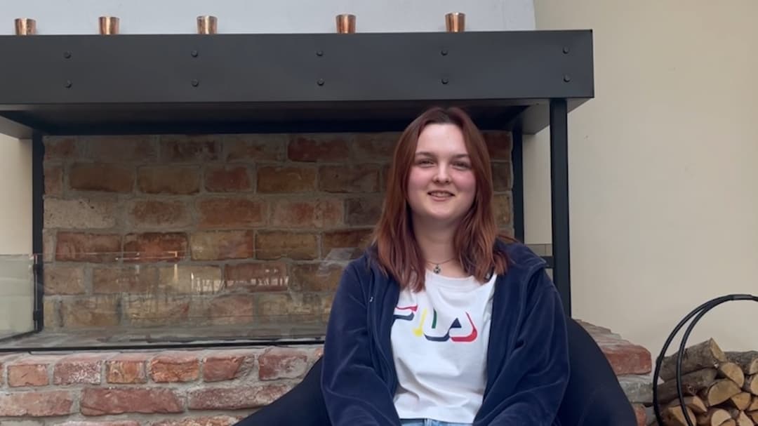 Kvinna sitter framför en eldstad, hon ler och tittar in i kameran