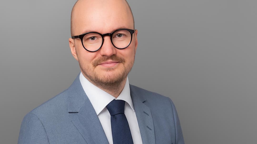 Man med glasögon och skägg, klädd i ljusgrå kavaj, vit skjorta och slips