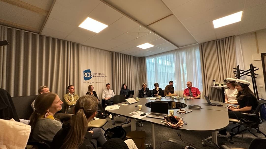 Fyra personer sitter runt ett ovalt konferensbord med laptops, papper och kaffekoppar. En skylt med texten "Risk and Crisis Research Centre" syns på väggen bakom dem.