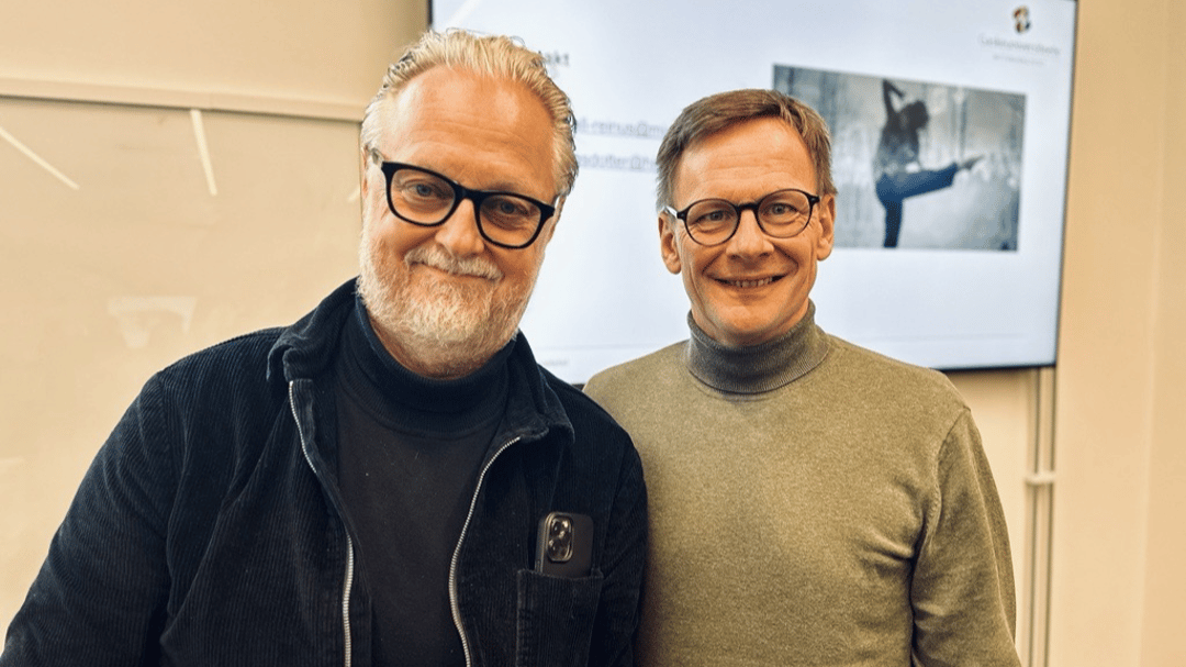 Två män i glasögon tittar in i kameran och ler. De står framför en skärm. 