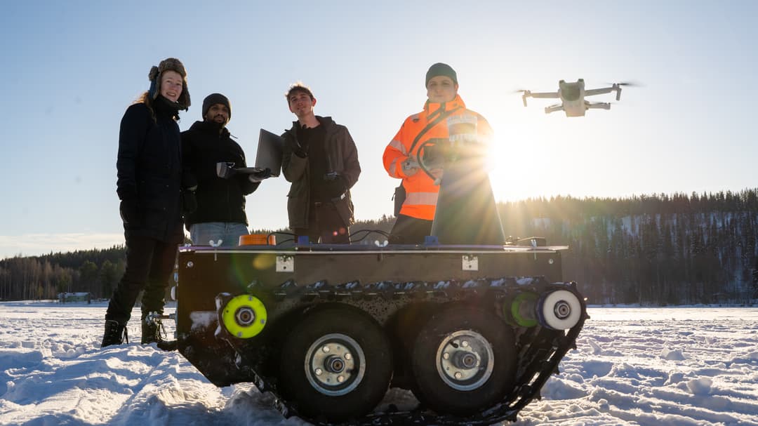 Fyra personer står i ett snöigt landskap. En drönare flyger bakom dem och framför dem står ett mindre terrängfordon.