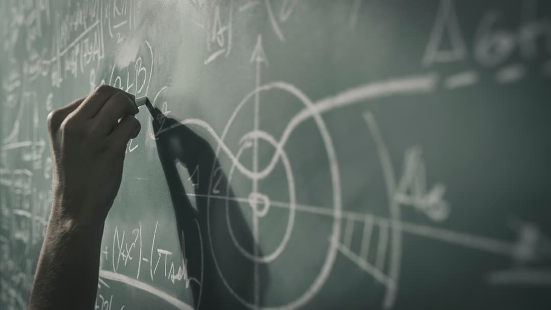  A hand holds a white chalk and writes formulas on a blackboard.