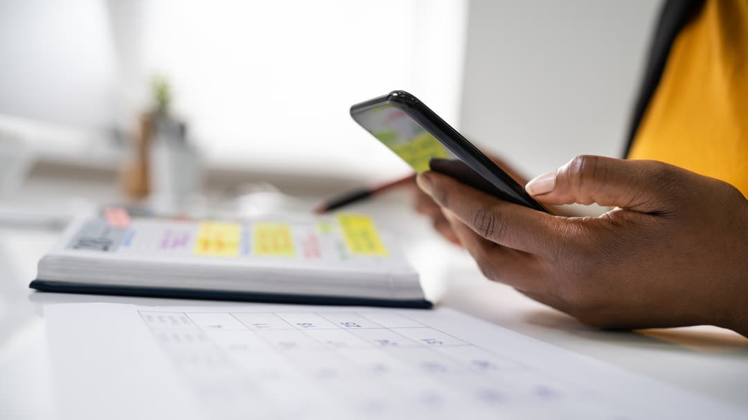 En hand håller i en mobiltelefon och en kalender ligger på ett bord.