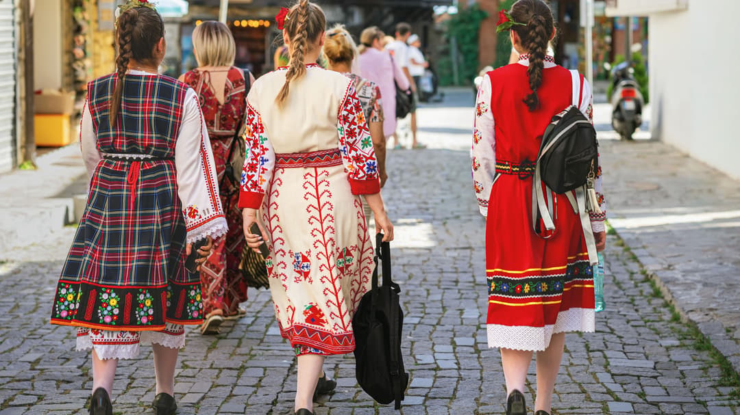 Tre kvinnor i traditionella bulgariska folkdräkter går på en gata i en stad. Dräkterna är färglada med starka färger och mönster. 