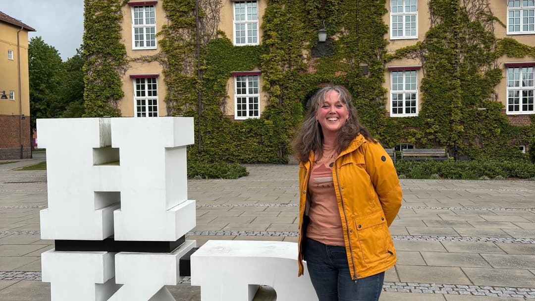 A woman in a yellow jacket is standing outside a beige building with green climbing plants. Next to her is a sculpture with the letters ‘HKR’.