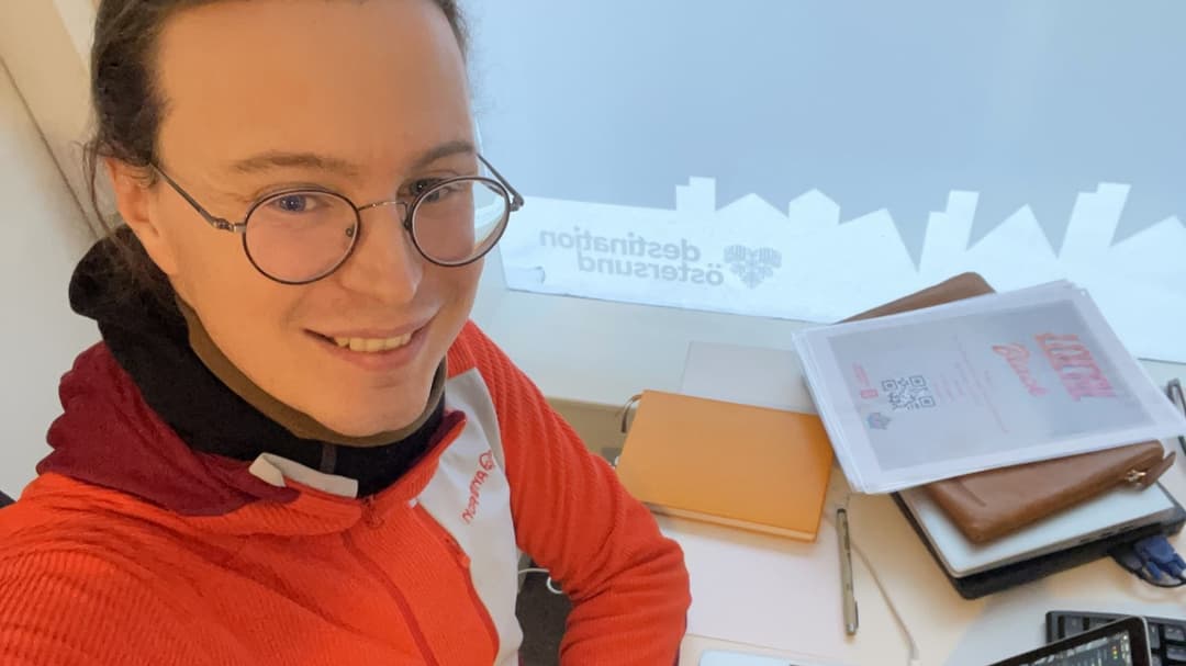 Ung man i glasögon och orange tröja som sitter med sin dator vid ett skrivbord  