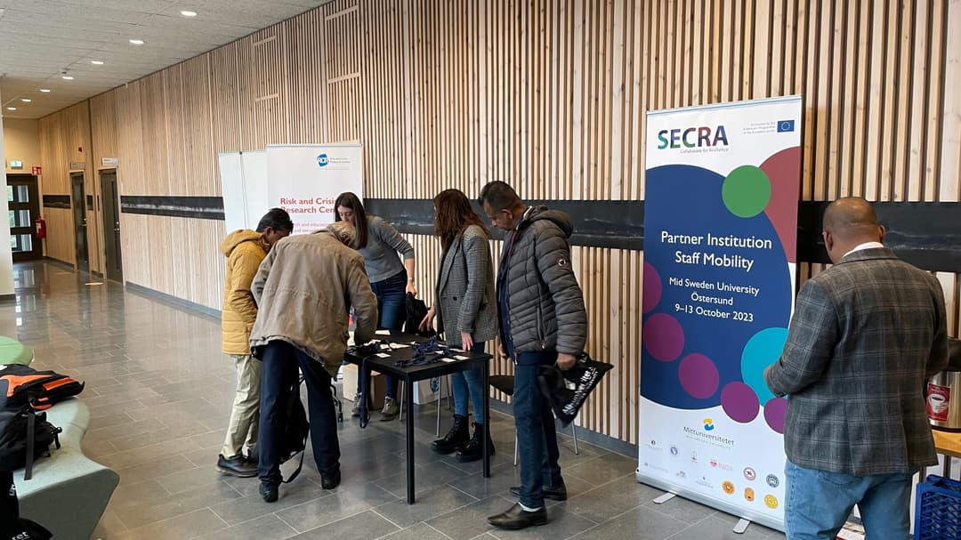 Några personer står vid ett bord. Bredvid bordet står en roll-up med texten "SECRA".