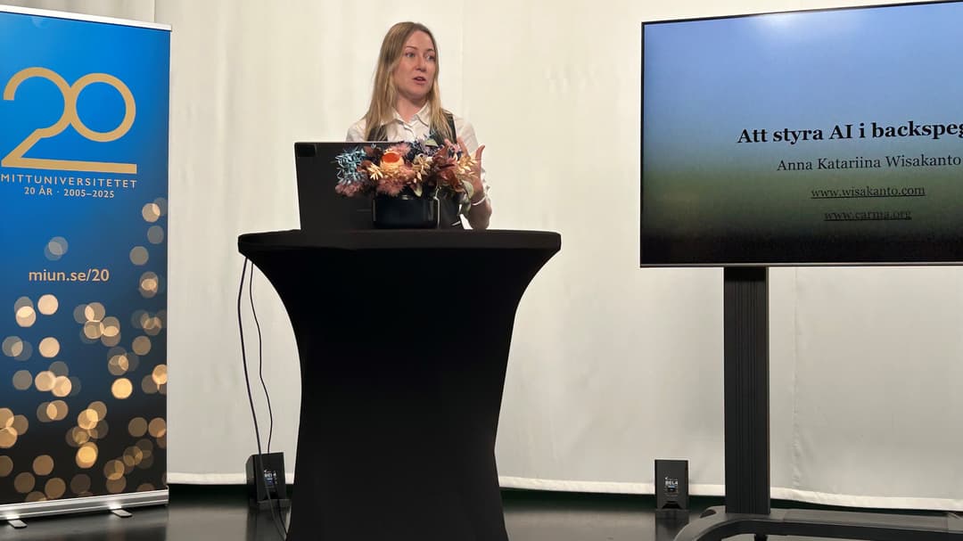 En blond kvinna står vid ett podium. I bakgrunden syns en stor tv-skärm med en presentation på. 
