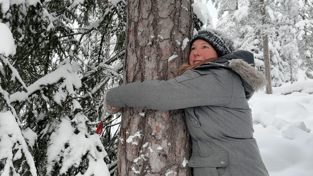 Kvinna kramar om trädstam i skogen, snö runt omkring
