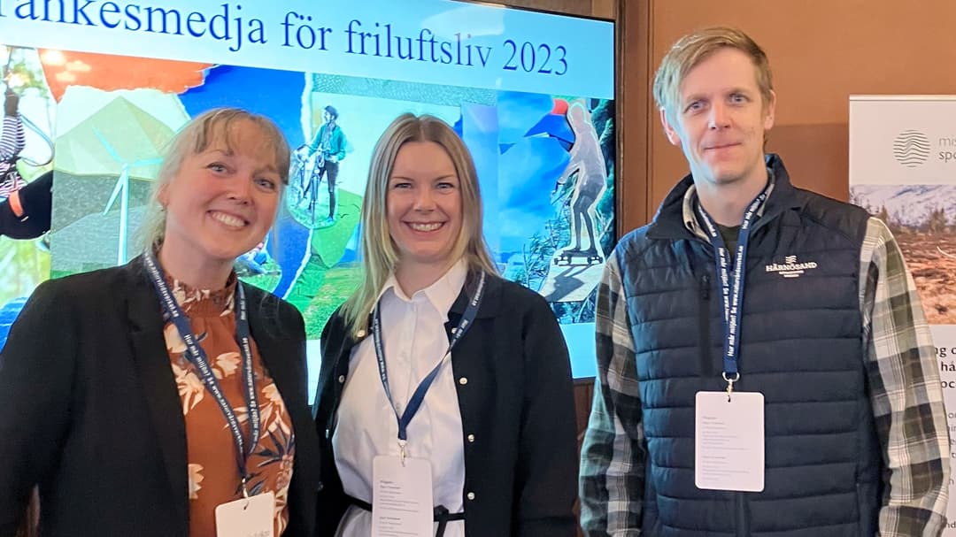 Två kvinnor och en man står i ett rum och tittar in i kameran. Bakom dem finns en skärm med en presentation på och de har namnskyltar hängande runt halsen 