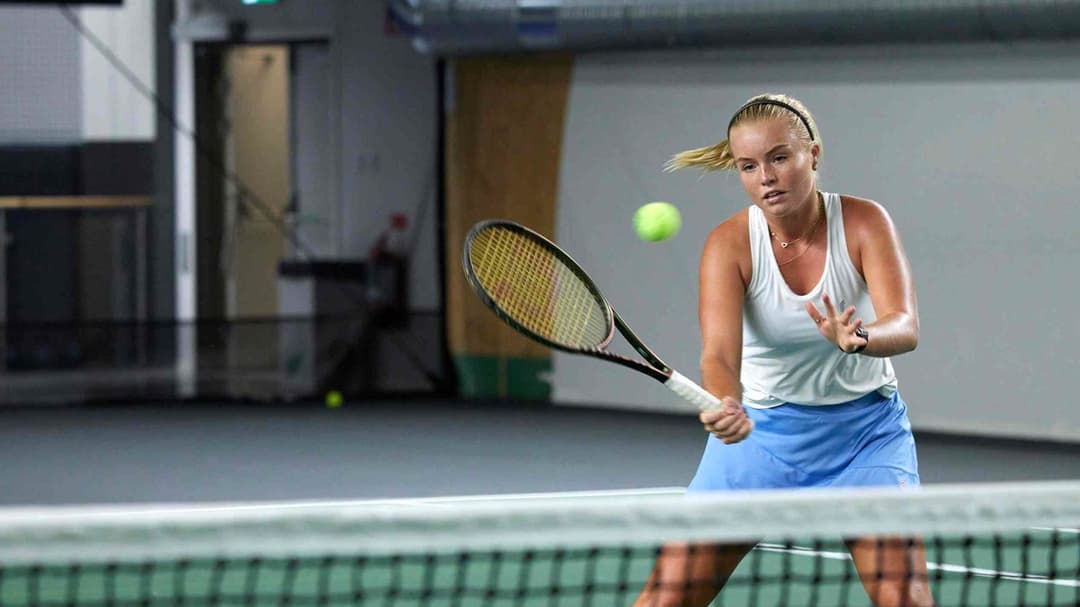 Kvinna som spelar tennis inomhus, hon står vid nät och ska till att slå en volley.