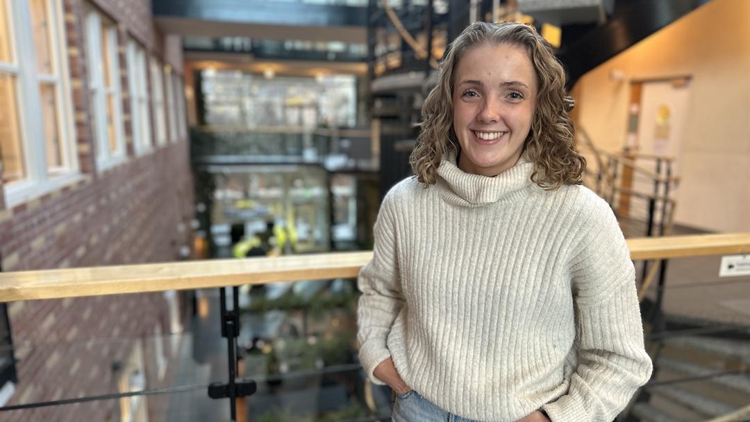 Ung kvinna med blont, lockigt, axellångt hår klädd i vit, stickad polotröja och ljusa jeans står i en ljus byggnad med många fönster, ett räcke och en trappa bakom sig. 