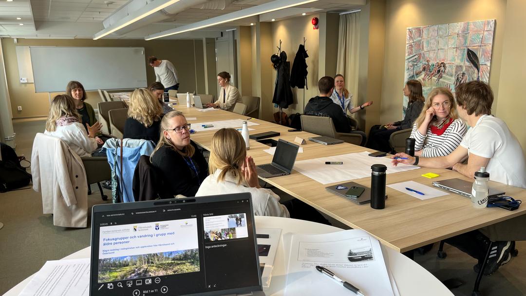 Ett tiotal personer sitter runt ett bord och pratar med varandra och tittar på en skärm med en presentation