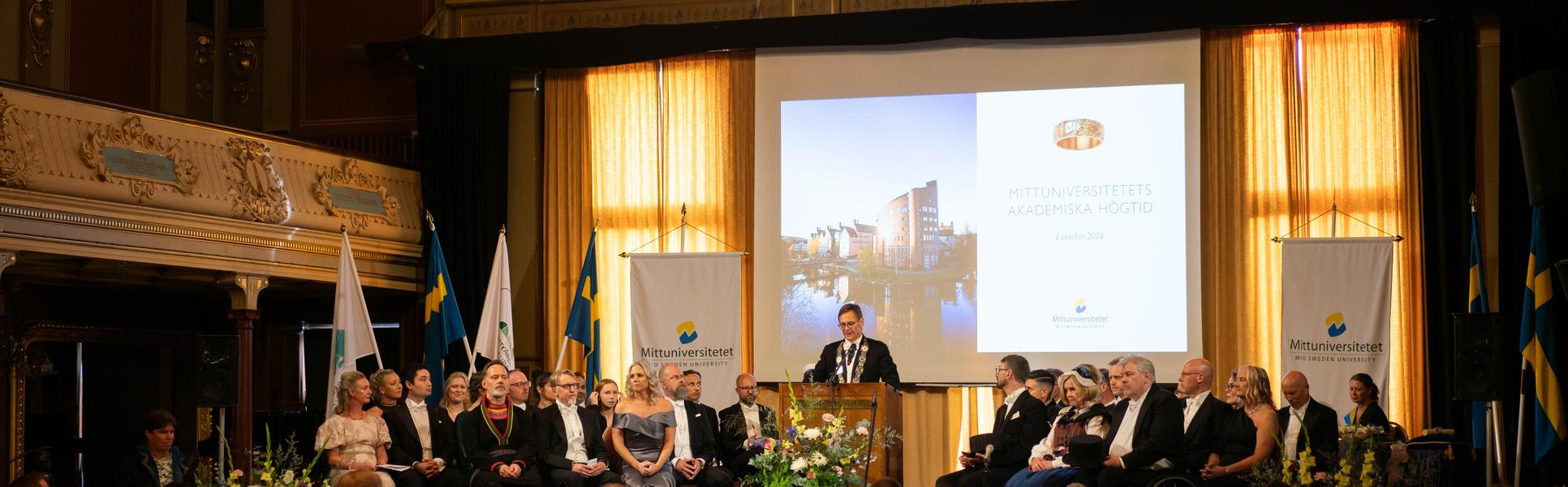 En finklädd man står i en talarstol och talar framför en publik.