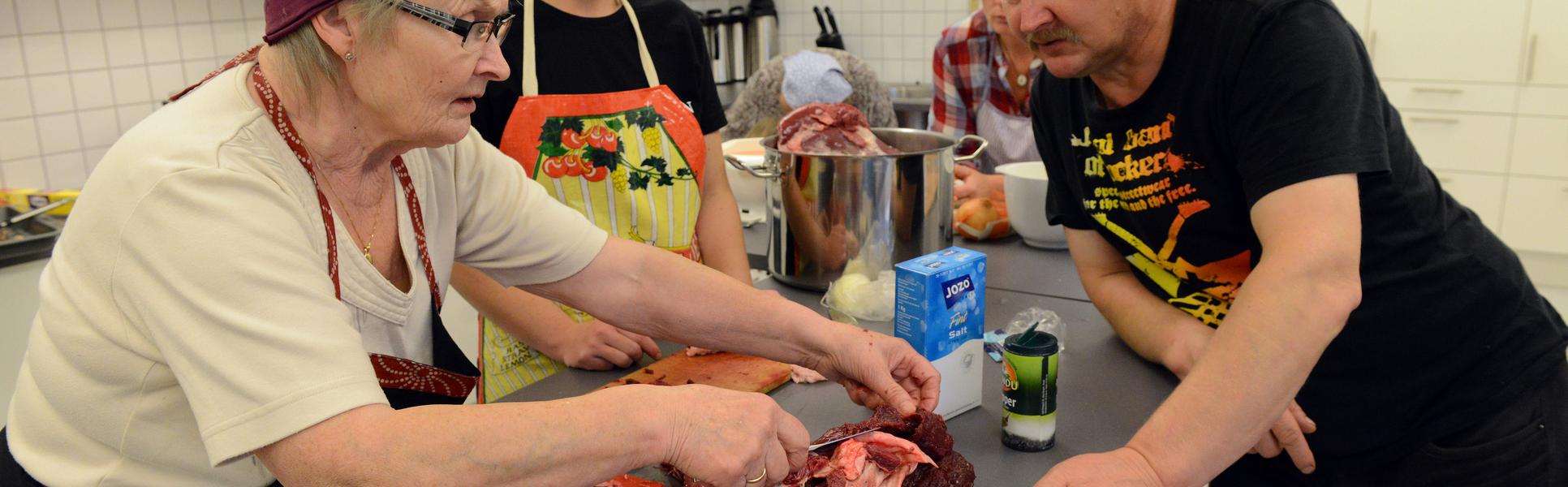 En äldre kvinna visar en ung kvinna och en man hur man tillagar renkött.