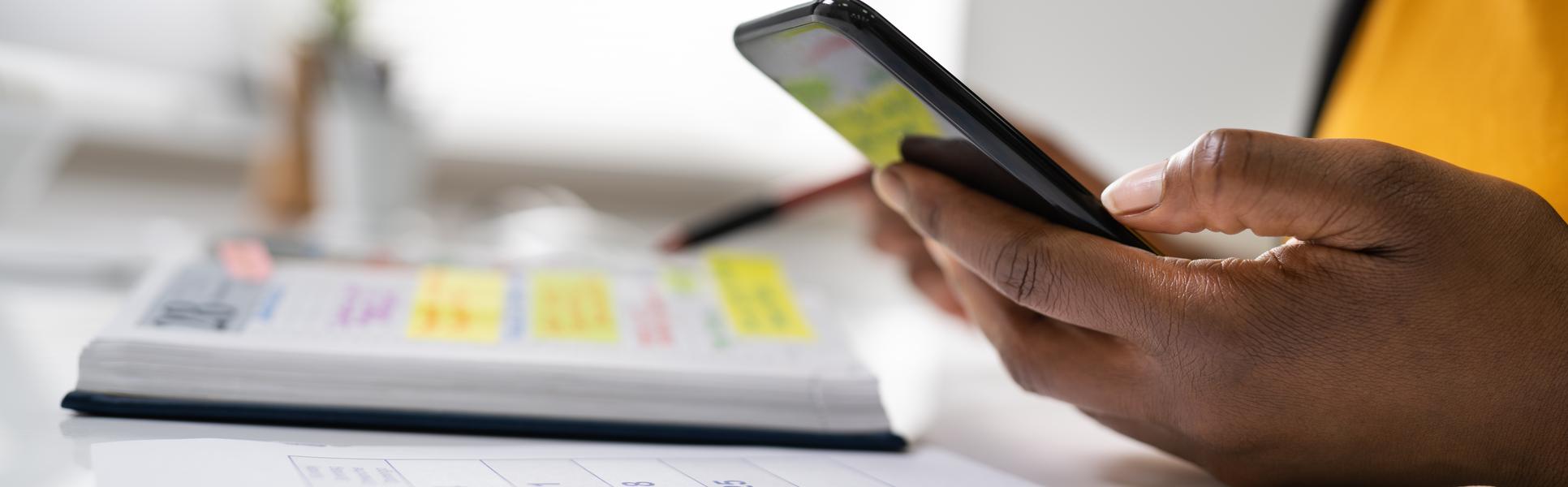En hand håller i en mobiltelefon och en kalender ligger på ett bord.
