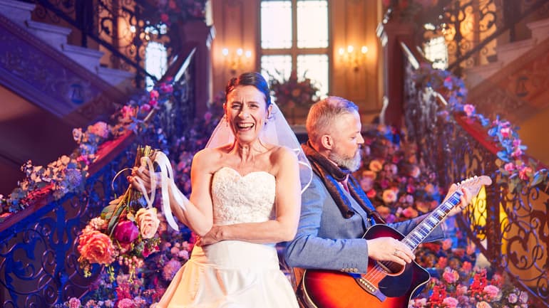 Kvinna i brudklänning, man i grå kostym med gitarr. Båda står framför en trappa fylld med blommor.