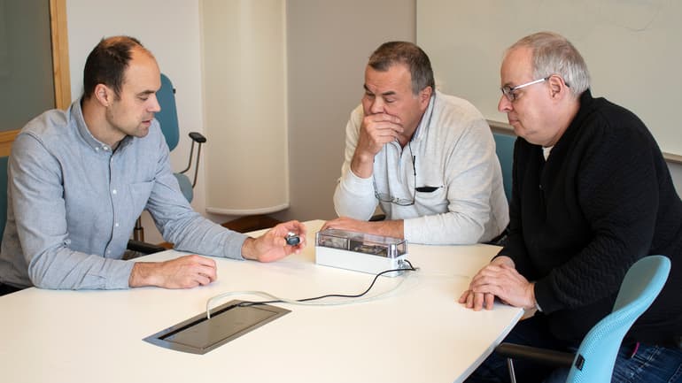 Tre män sitter vid ett bord och diskuterar en prototyp som ligger på bordet.