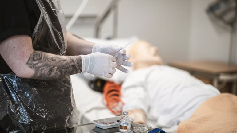 En man med skägg är i en sjukhusmiljö och övar med en spruta på en patientdocka.
