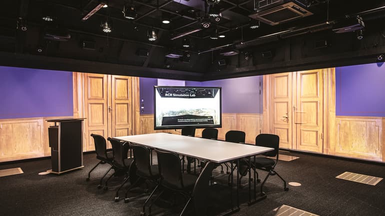 A large room with black floor and ceiling. Images from a grand meeting room are projected on the walls. The room is furnished with a large table and eight chairs, a computer screen and a lectern. There are lots of technical equipment in the ceiling.
