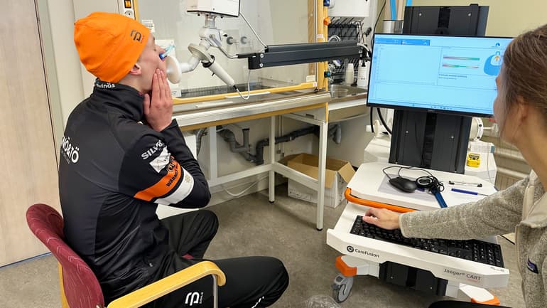 Ung man med toppluva blåser i ett munstycke, testledare övervakar på bildskärm.