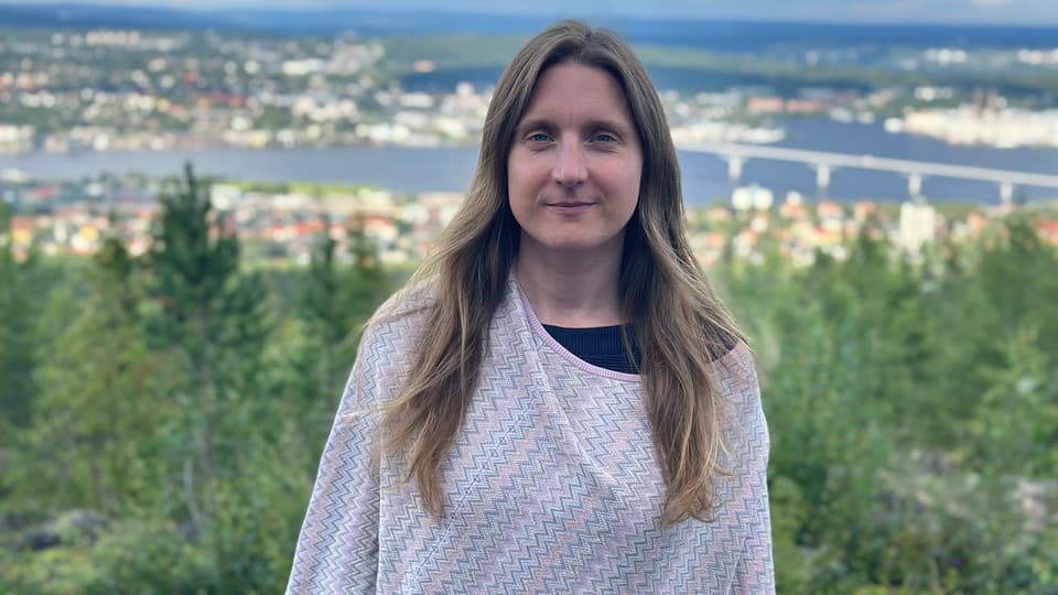 Kvinna med långt mörkt hår, en mönstrad överdel i pastellfärger, i bakgrunden se man Sundsvall.