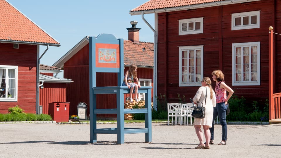 En flicka sitter på stor stol utanför tre röda hus och två kvinnor står nedanför och tittar på flickan.