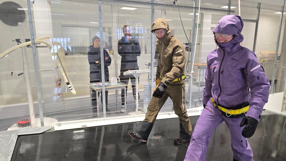 Two people in colorful clothes walk in a glass tunnel while two people watch.