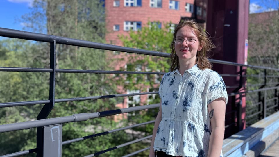 A student is smiling. In the background the campus is visable. 