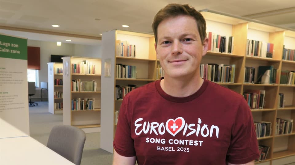 Ung kortklippt man i mörkröd t-shirt med Eurovision-tryck, sittandes vid ett bord i ett bibliotek med böcker i bakgrunden