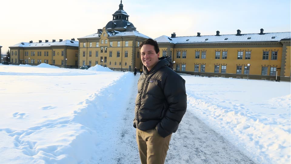Man in a winter jacket is standing infront of yellow buildings at Mittuniversitetets campus in Östersund