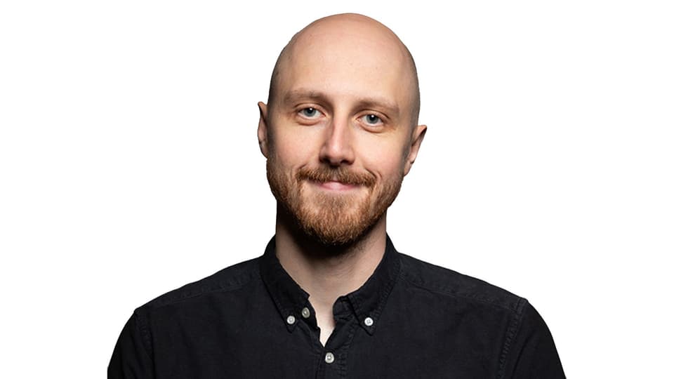 Man med skägg och mustasch ler mot kameran, klädd i svart skjorta, vitt bakgrund.