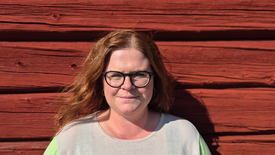 En kvinna med brunt hår och glasögon mot en röd trävägg