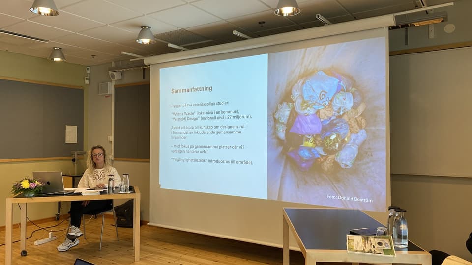 En kvinna i lockigt ljust hår sitter vid ett bord och håller en presentation som visas på en stor duk.
