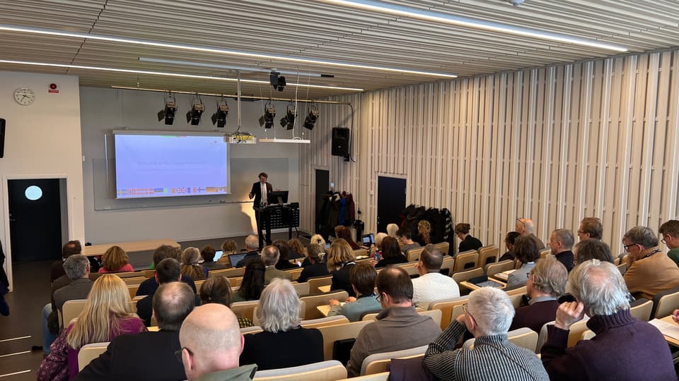 Åhörare sitter i föreläsningssal med ryggen mot kameran