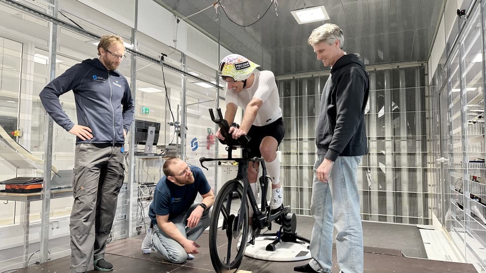 Tre män står och tittar på en fjärde man som sitter på en cykel inomhus.