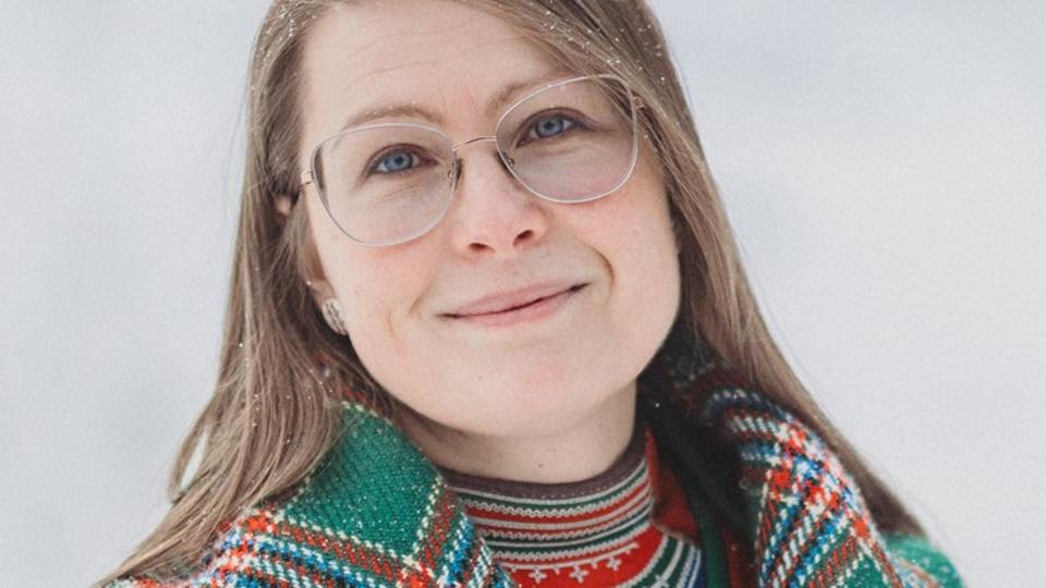 Woman with glasses and dark blonde hair with a chequered shawl over her shoulders looks at the camera.