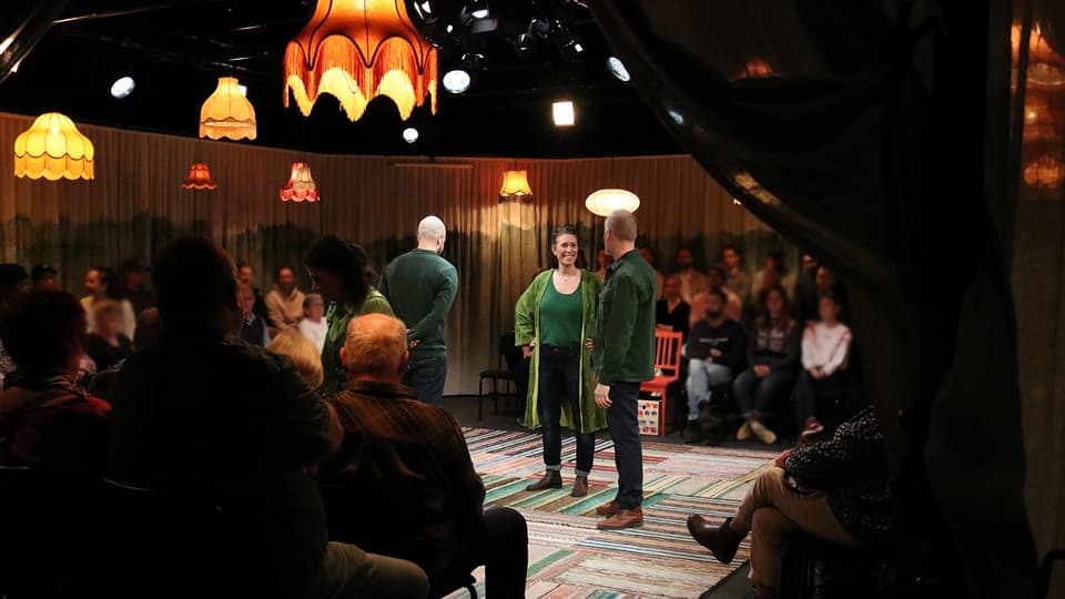 En teaterscen med tre personer i gröna tröjor på och publik runtomkring. 