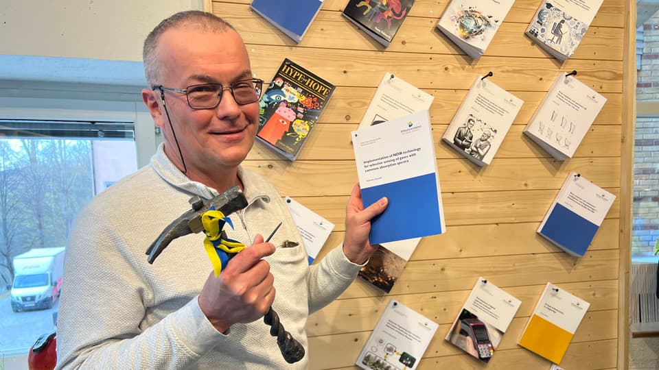 En man har en avhandling i ena handen och en hammare i den andra handen. Han står framför en vägg med uppspikade avhandlingar.